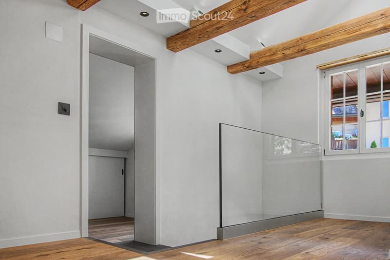 Empty room with wooden flooring, white walls, exposed wooden beams, a glass partition, and a window showing a view of another room.