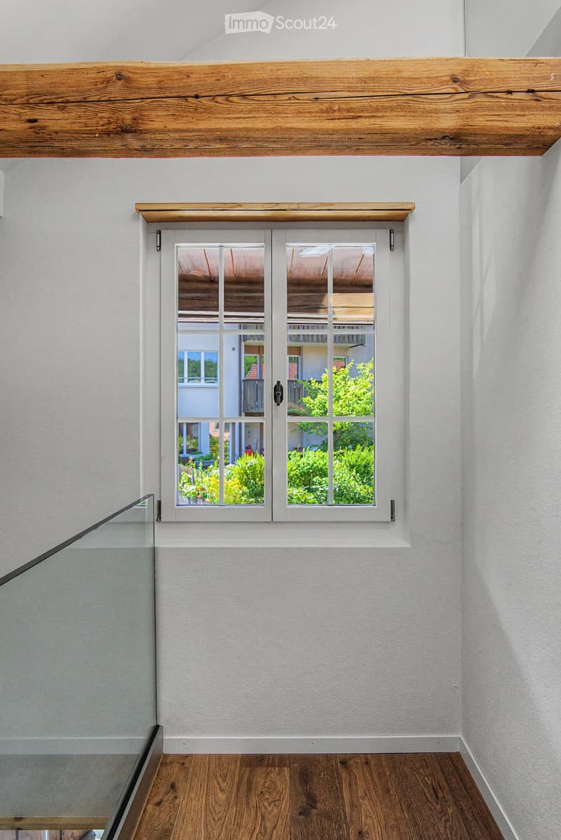 Modern hallway with white walls, wooden floor, and a large window offering a view of the garden and neighboring house.