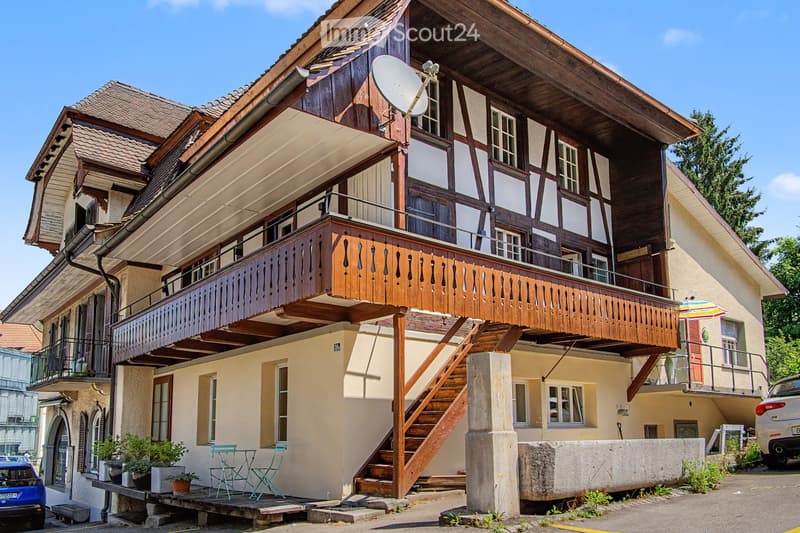 Traditional two-story house with wooden balcony, satellite dish, and small garden area with chairs and tables.