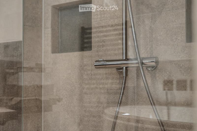 Modern showerhead with dual spray options mounted on beige tiled wall, glass shower enclosure visible.