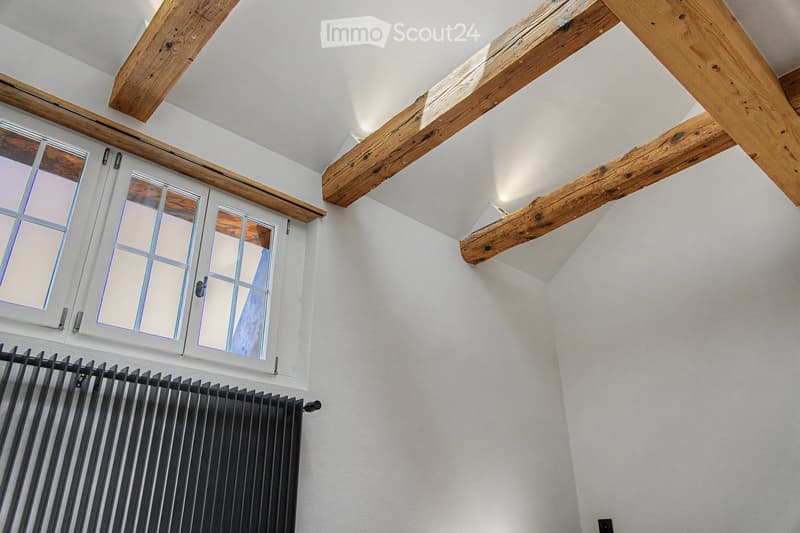 A hallway with exposed wooden beams, white walls, and a window with multiple panes. A black radiator is visible below the window.