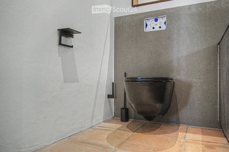 Modern bathroom with a dark grey toilet, a toilet brush holder, and a toilet paper holder mounted on the wall. The walls are painted in a two-tone design with white lower half and grey upper half, and the floor is tiled in warm beige tiles.