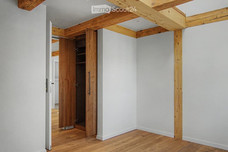 Empty room with exposed wooden beams on the ceiling, light grey walls, and wooden flooring. A wooden closet door is open, revealing empty shelves.