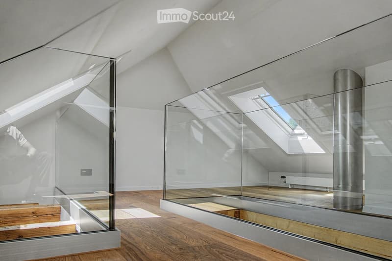 Bright, minimalist attic space with wooden flooring, white walls, and large skylights. Glass partitions separate areas, and a metal column is visible.