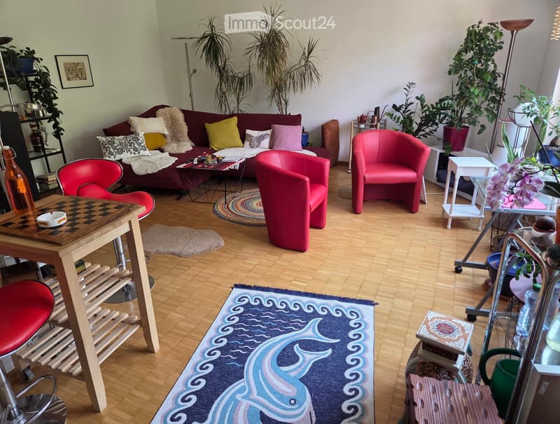 A cozy living room with a red sofa, colorful cushions, a wooden coffee table, red chairs, and various plants. The room features wooden flooring and multiple rugs.