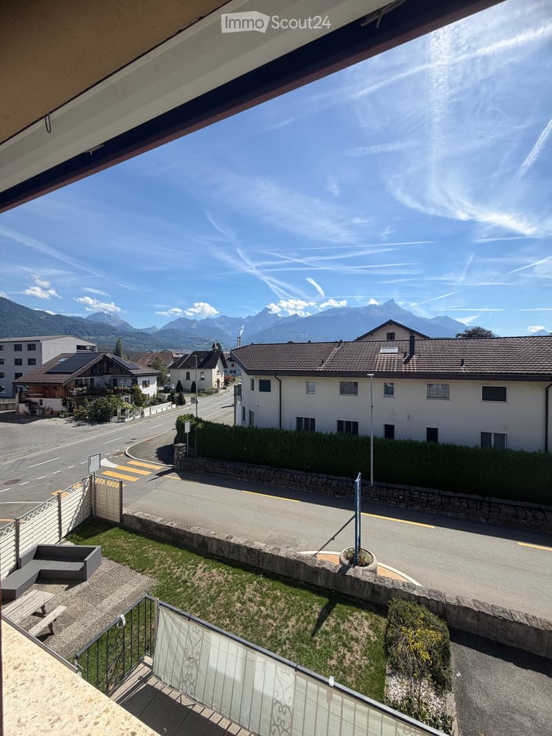 Balcony overlooking a street with houses, a stone wall, a small grassy area, and a view of mountains in the distance.