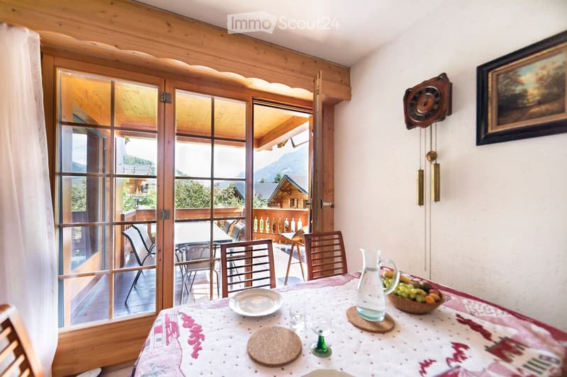 A cozy dining area with a table set for a meal, chairs, and a view of a wooden balcony and mountain landscape through large glass doors.