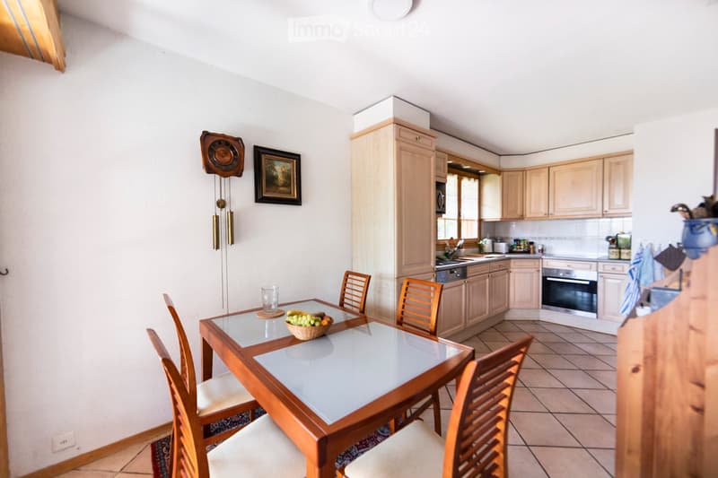 Bright kitchen with light wooden cabinets, a dining table with chairs, and a bowl of fruit on the table. The kitchen features a sink, oven, and dishwasher, with a framed picture and a wall-mounted clock on the wall.