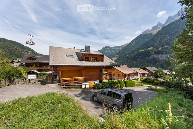 A large wooden chalet with a gray roof, surrounded by lush greenery and mountains, with a cable car passing overhead.