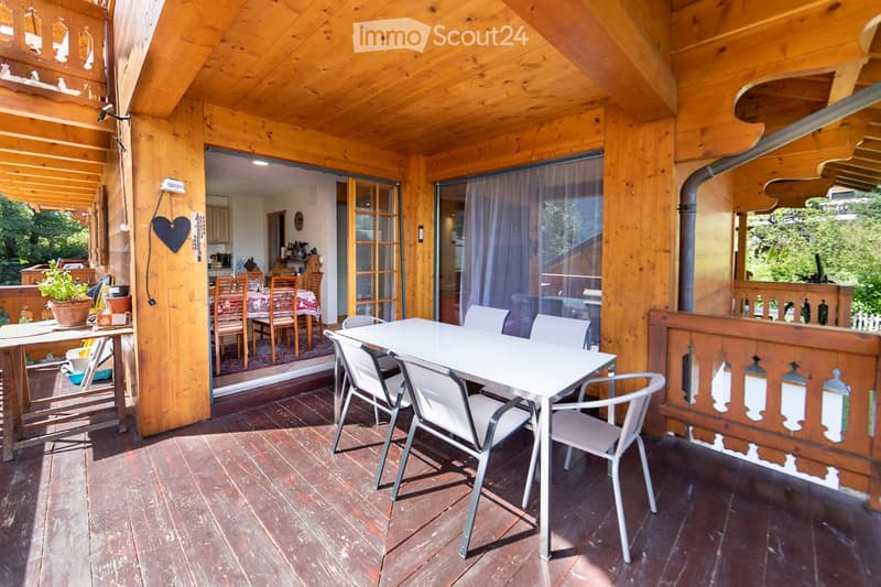 Wooden balcony with a white table and chairs, overlooking a dining area inside the house.