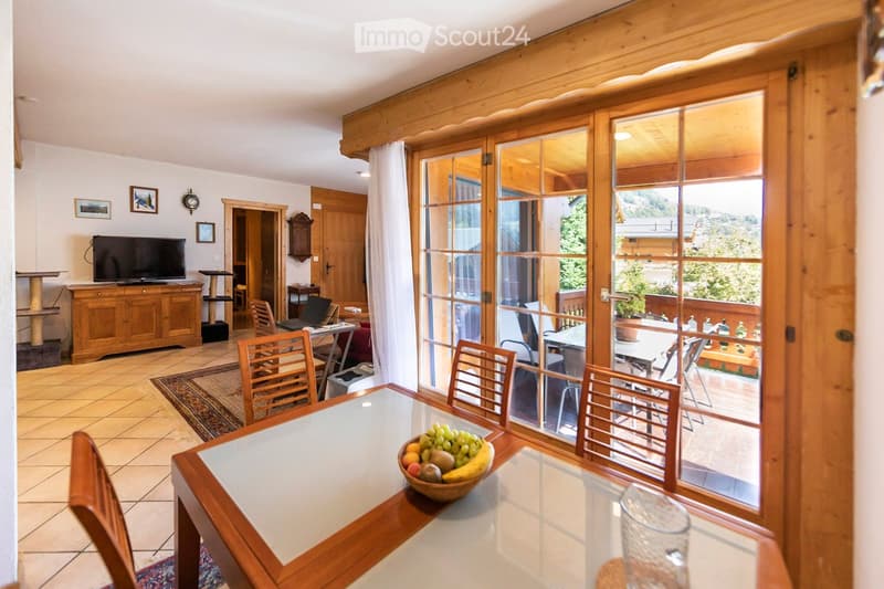 Bright living and dining area with large glass doors leading to a balcony, wooden furniture, a dining table with chairs, a bowl of fruit, a laptop, a cat tree, and a television on a wooden cabinet.
