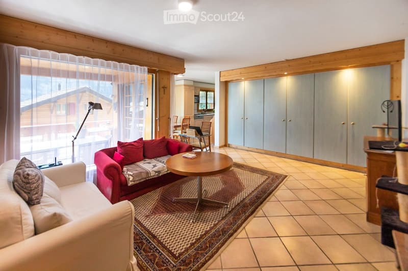A cozy living room with a red sofa, a round coffee table, a patterned rug, and a view of a wooden house through large windows.