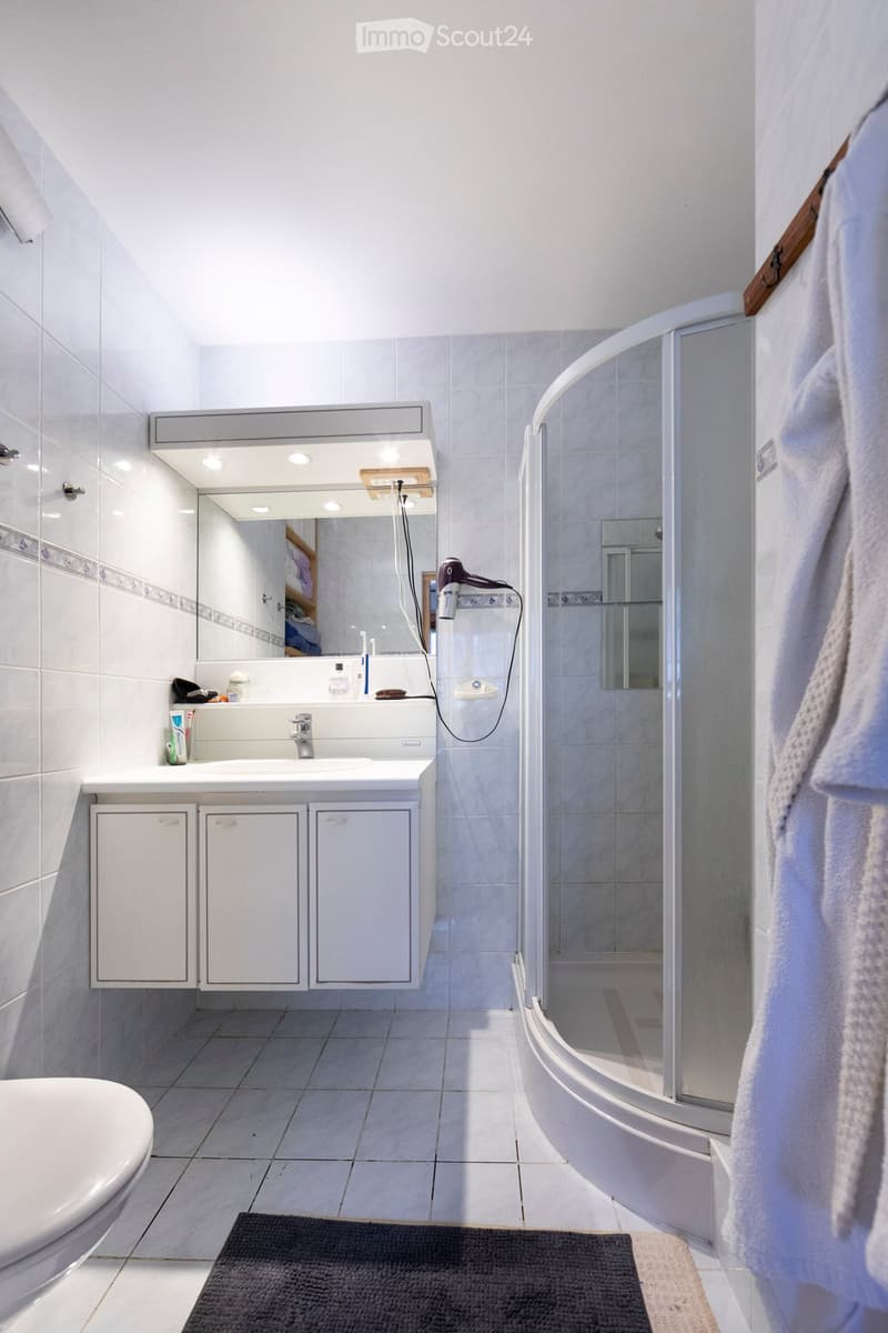 Modern bathroom with white tiles, a large mirror, a sink with a cabinet underneath, a toilet, a shower with a glass enclosure, and a towel hanging on the side.