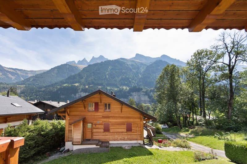 A wooden chalet with a sloped roof and red shutters, situated in a lush green area with a scenic mountain backdrop.