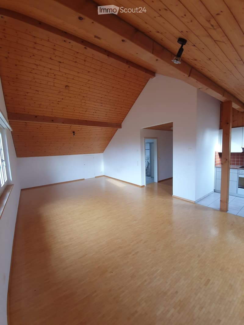 Empty attic space with wooden floors and exposed wooden beams on the ceiling. Natural light streams in from a window on the left, and there are visible doorways leading to other rooms.