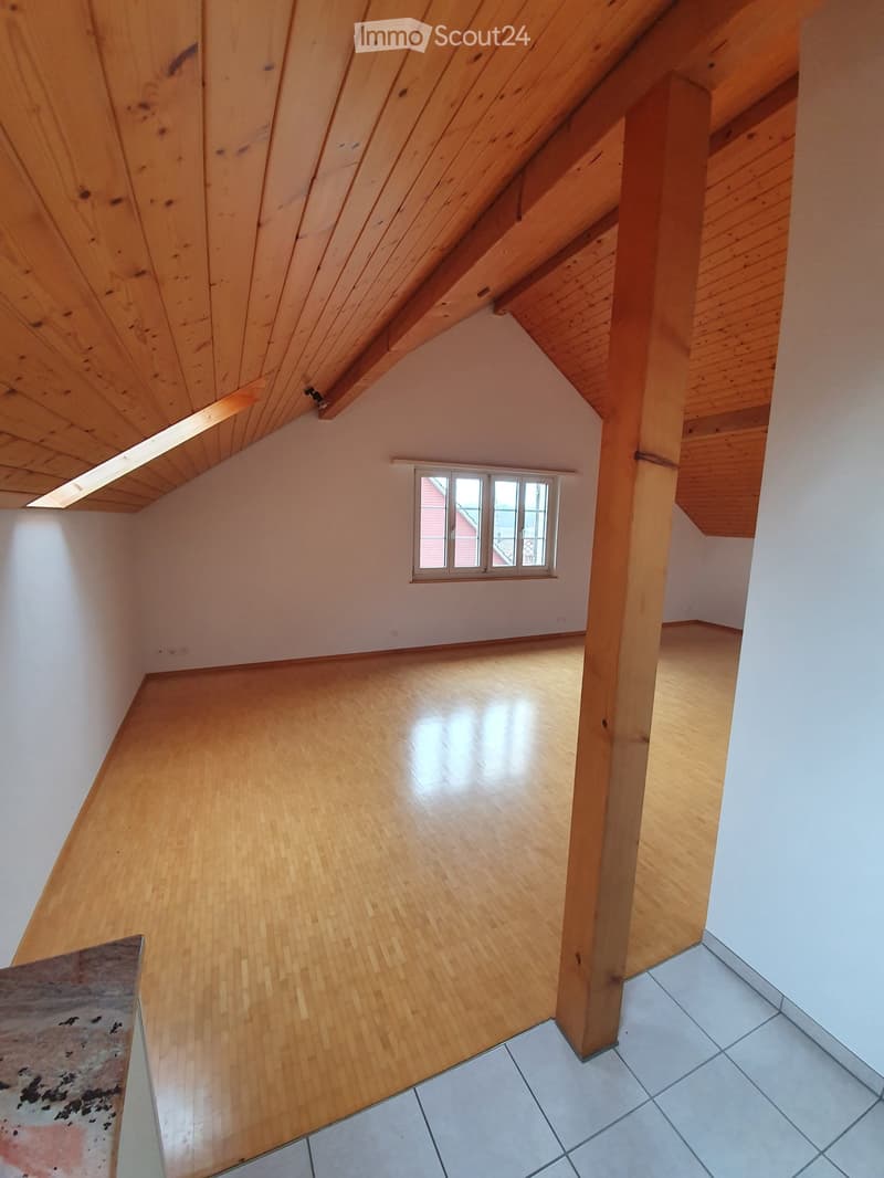 Empty attic room with wooden slanted ceilings, light wooden flooring, and a small window letting in natural light.