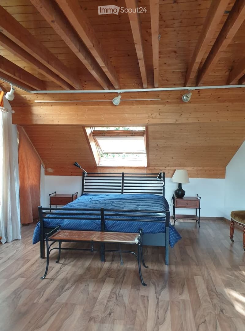 Cozy attic bedroom with wooden beams, exposed ceiling, large skylight, and modern furnishings including a bed, nightstands, and a bench.