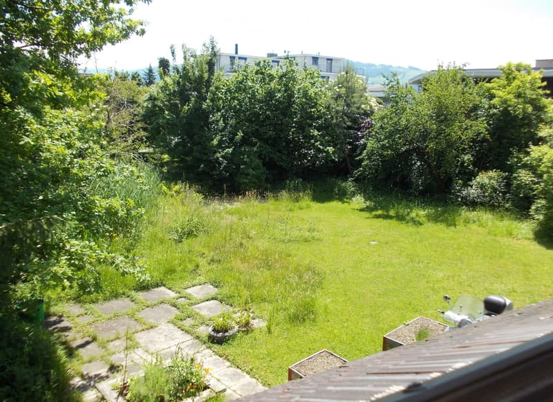 Lush green garden with a stone pathway leading to a grassy area, surrounded by trees and shrubs. A scooter is parked on the right side, and a small planter is visible near the pathway.