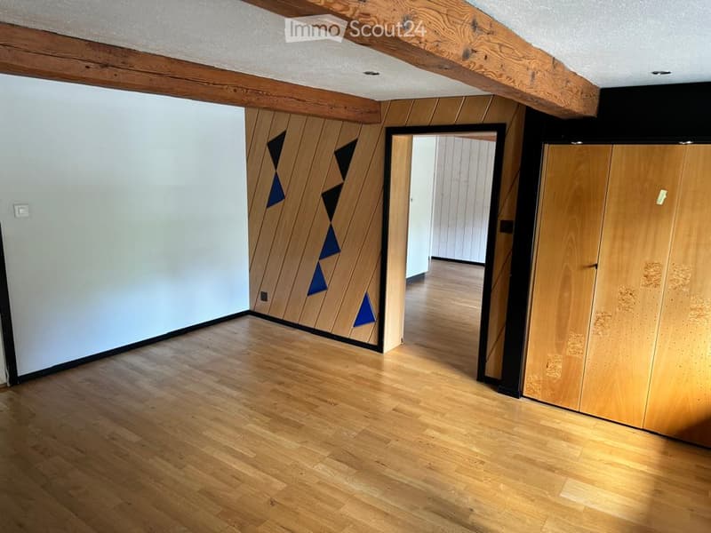 Empty room with wooden flooring, white walls, exposed wooden beams on the ceiling, and a doorway leading to another room.
