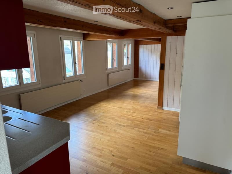 Empty room with wooden flooring, exposed wooden beams on the ceiling, and large windows with white frames. The walls are painted in a neutral tone, and there is a radiator below one of the windows.