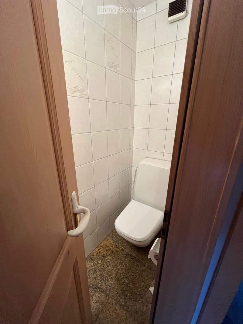 Small bathroom with white tiled walls, a toilet, and a toilet paper roll on the floor. The door is slightly open, revealing a wooden frame and a white handle.