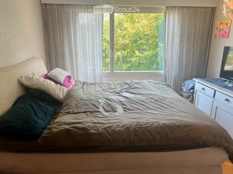 A well-lit bedroom with a large bed, nightstand, television, and large windows with curtains.
