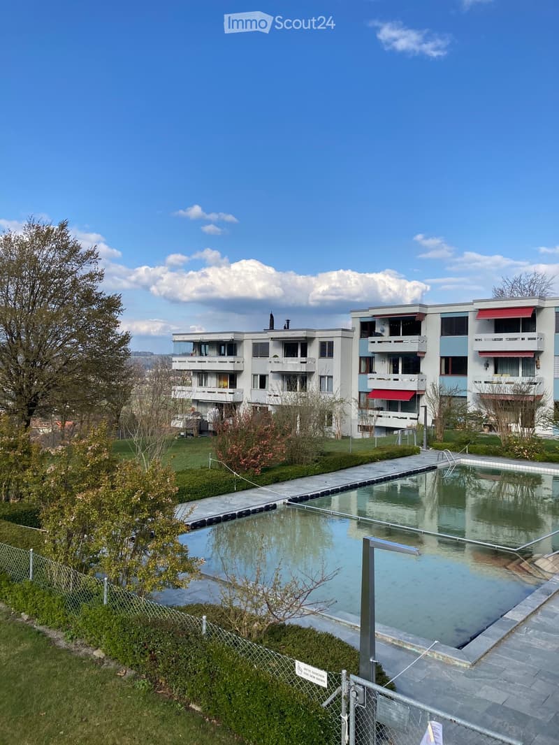 Modern residential complex with a large swimming pool in the foreground, surrounded by greenery and pathways.