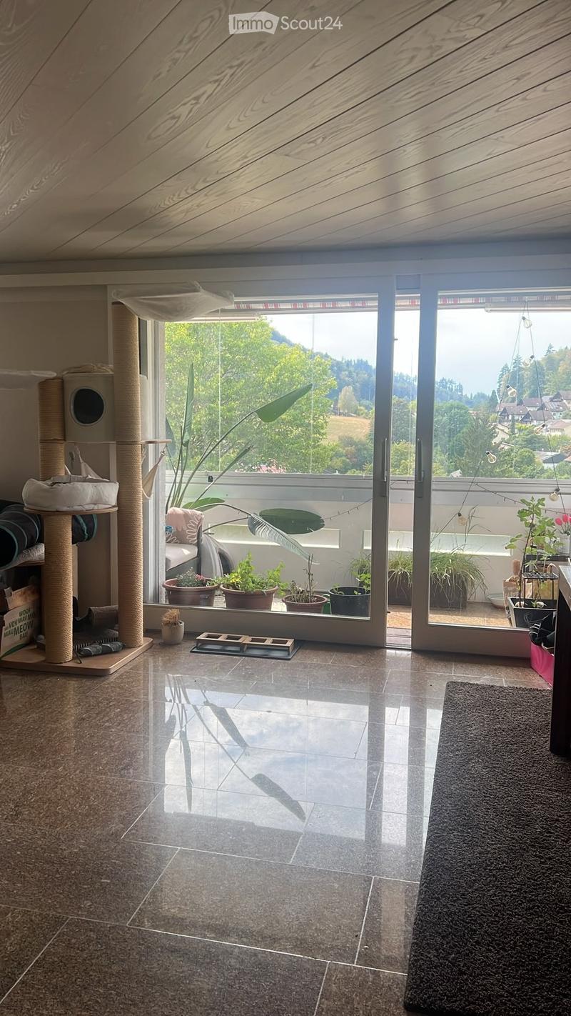 A modern, well-lit room with a large sliding glass door leading to a balcony. The room features a cat tree, a rug, a small table with a chair, and several potted plants. The floor is tiled, and the ceiling has wooden paneling.