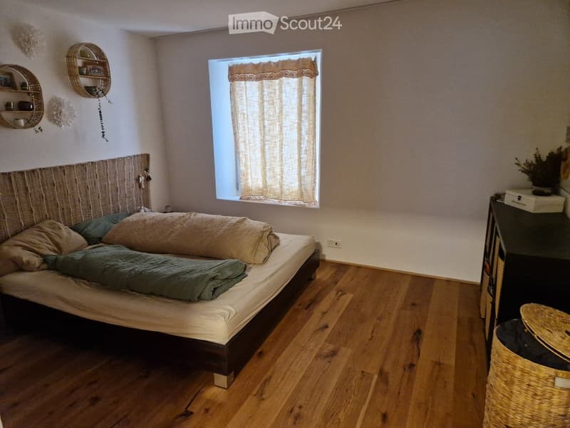 Cozy bedroom with wooden flooring, a large bed with a woven headboard, and a nightstand with a lamp. The room features a window with sheer curtains and decorative wall shelves.