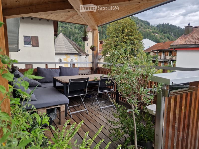 Wooden balcony with a sofa, table, chairs, and potted plants, overlooking a scenic town with historic buildings.