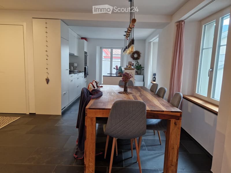 Bright, modern dining area with a long wooden dining table surrounded by grey upholstered chairs. The space features a sleek kitchen area with white cabinets, an oven, and a faucet. Natural light streams in through large windows with pink curtains, and de