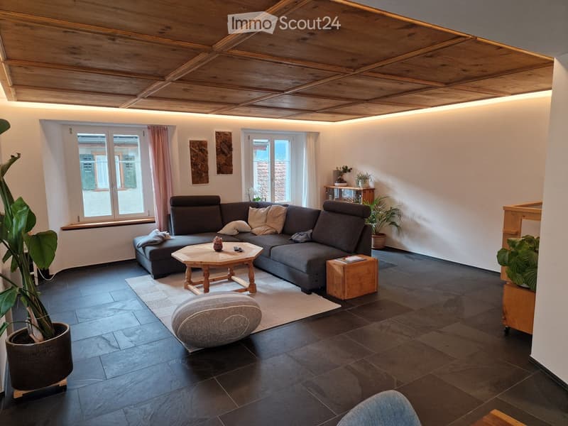 Modern living room with dark grey sectional sofa, wooden coffee table, ottoman, side table, and potted plants. The room features exposed wooden ceiling beams and large windows with sheer curtains.