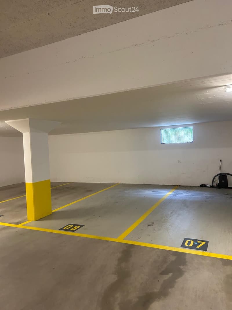 Empty underground parking space with marked parking spots, white walls, and a single window at the far end.