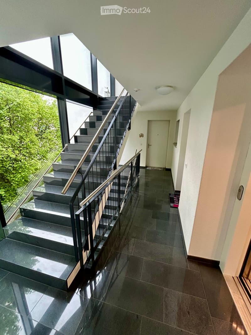 Modern hallway with polished tile flooring, a staircase with metal railing, and large windows offering a view of greenery.