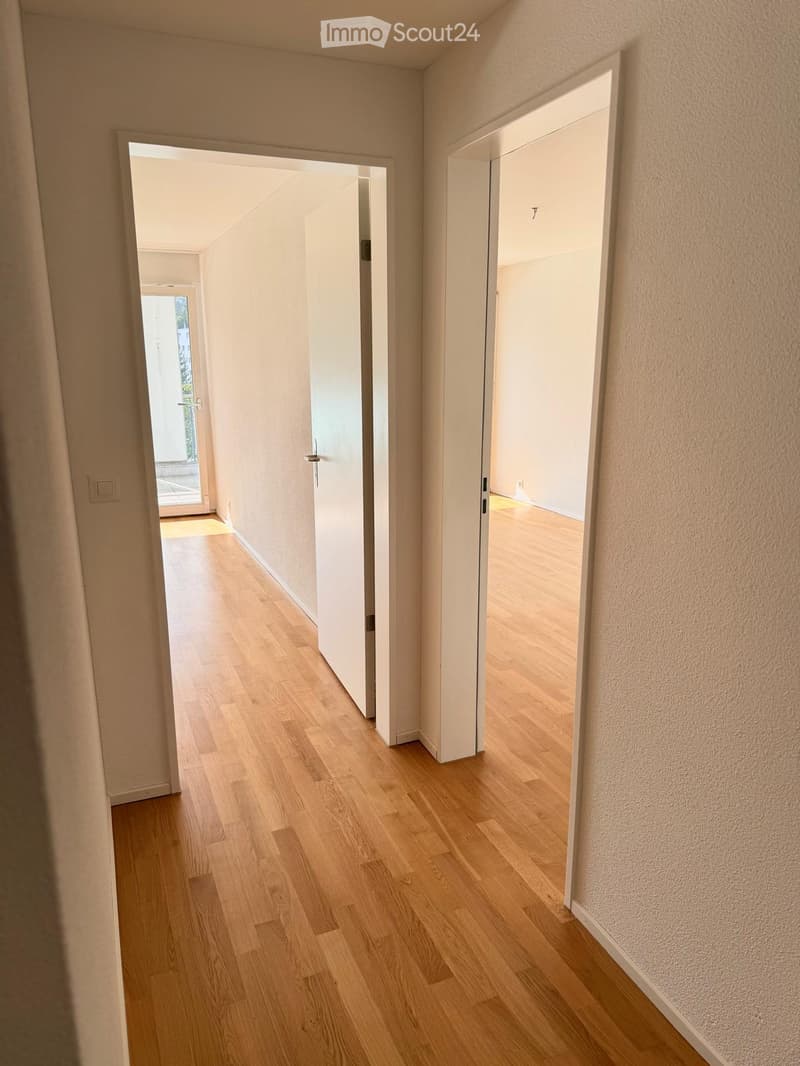 Empty hallway with light wooden flooring and white walls, featuring two open doors leading to adjacent rooms.