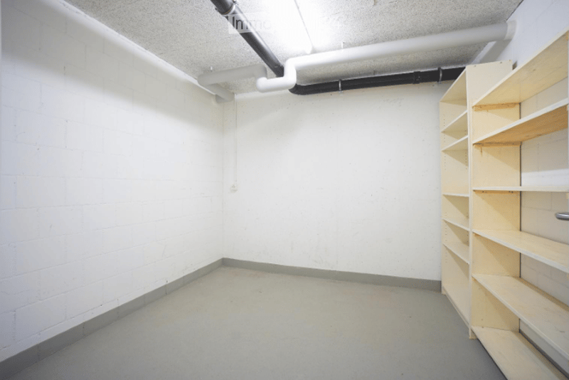 Empty room with white brick walls, grey concrete floor, and exposed pipes on the ceiling. A wooden shelving unit is present but no furniture or personal items.