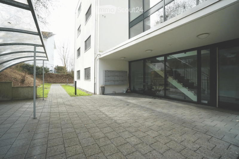 A paved outdoor corridor leading to a modern building entrance with glass doors and a staircase visible inside. The area is clean and well-maintained with a covered walkway on one side.
