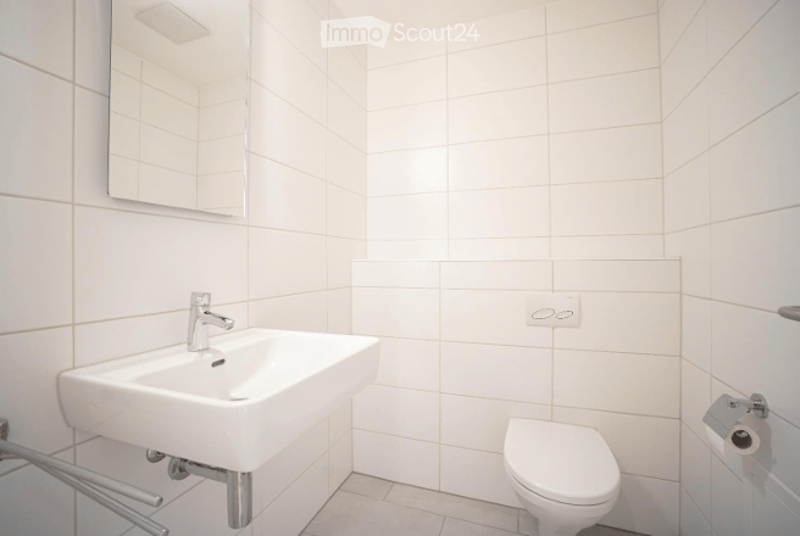 Modern bathroom with white tiled walls and floor, featuring a white sink with a chrome faucet, a mirror above it, and a white toilet with a toilet paper holder.
