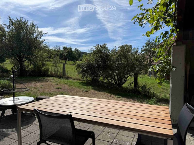 Outdoor patio with wooden table, chairs, and a view of a green landscape with trees and hills.