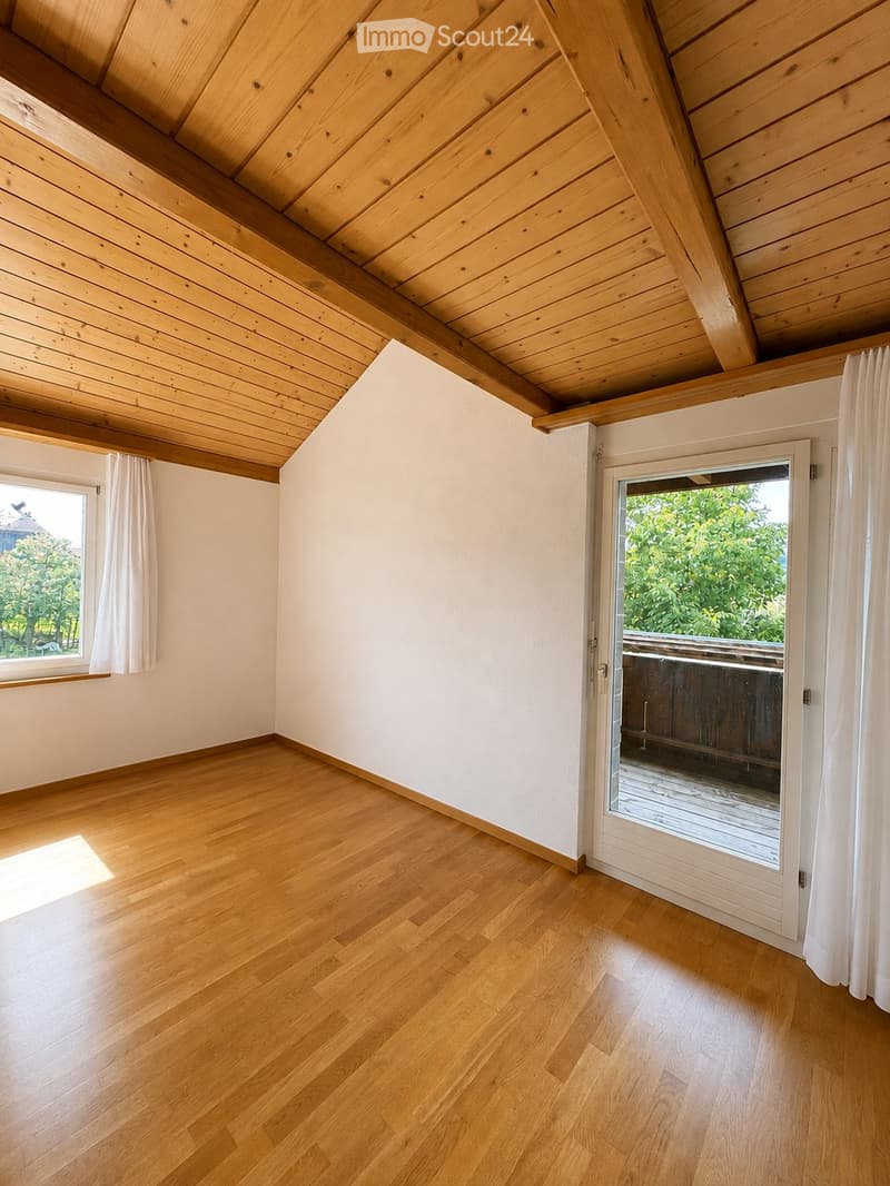 Empty room with wooden flooring, white walls, and a slanted wooden ceiling. Features a large window with white curtains and a sliding glass door leading to a balcony.