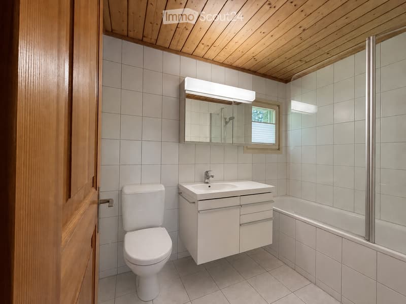 Modern bathroom with white tiles, a toilet, a sink with a mirror, and a bathtub. The ceiling is wooden, and there is a window above the bathtub.