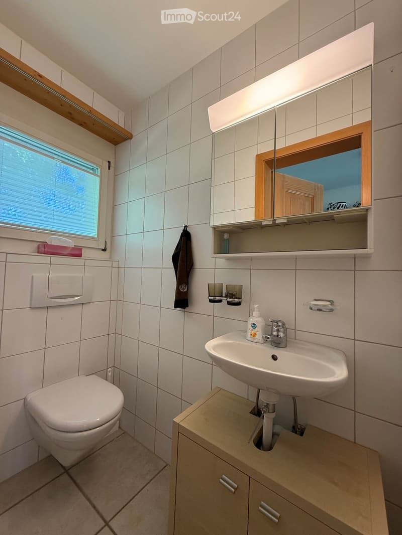 Modern bathroom with white tiles, a sink with a modern faucet, a toilet, a large mirror with built-in lighting, a wooden cabinet, and a towel hanging on the wall.