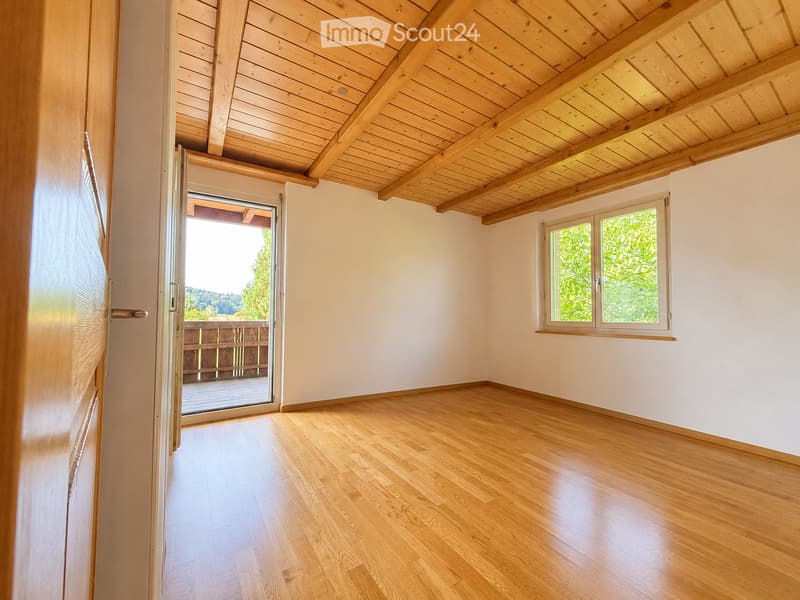 Empty room with wooden flooring, white walls, exposed wooden ceiling beams, large window, and sliding glass door leading to a balcony.