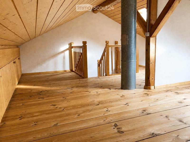 Spacious hallway with wooden flooring, exposed wooden beams, and a central column. Features a wooden staircase with a railing leading to an upper level.