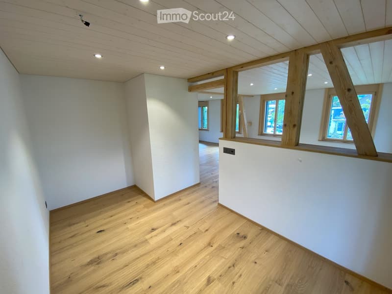 Empty room with light wooden flooring, white walls, and recessed ceiling lights. The room features large windows with wooden frames and a visible open doorway leading to another space.