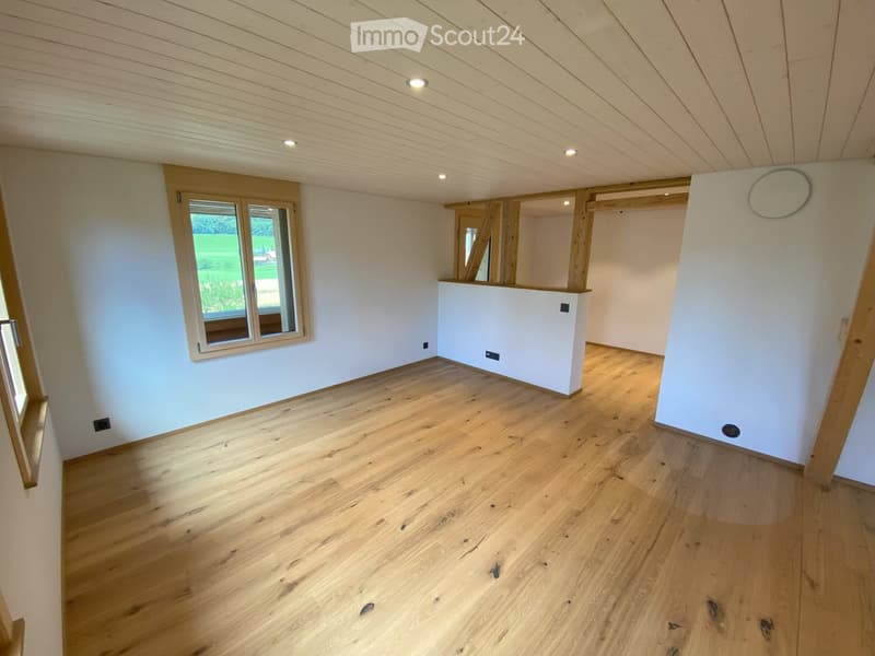 Empty room with light wooden flooring, white walls, and large windows offering a view of a green outdoor area. The ceiling has exposed wooden beams and recessed lighting.