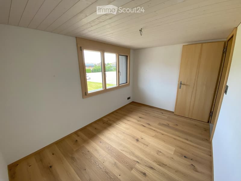 Empty room with light wooden flooring, white walls, and a large window with wooden frame. A wooden door is visible on the right side.