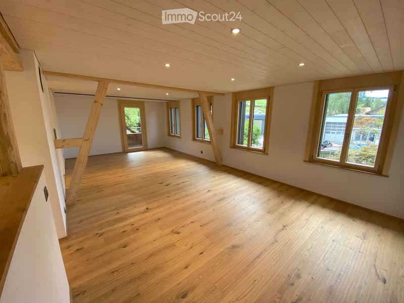 Empty room with wooden flooring, white walls, and large windows allowing natural light. Exposed wooden beams on the ceiling and a partially visible doorway leading to another space.
