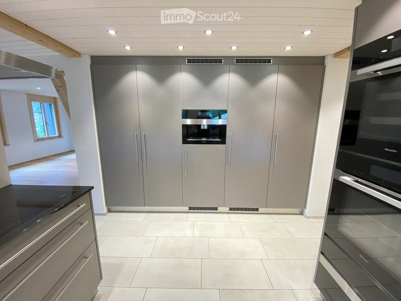 Modern kitchen with sleek grey cabinets, a black countertop, and built-in ovens. The space features recessed lighting and a tiled floor.