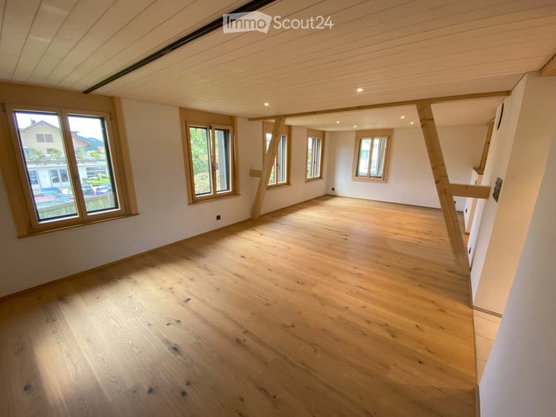 Empty room with wooden flooring, white walls, and multiple windows allowing natural light. The ceiling has exposed wooden beams and recessed lighting.